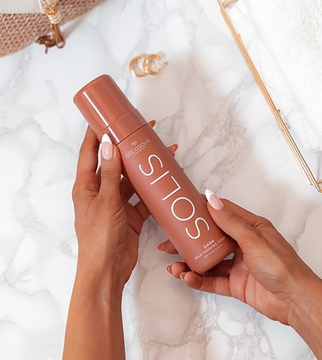 Two tan hands holding the COCOSOLIS SOLIS Self-Tanning Foam Bottle above a granite counter